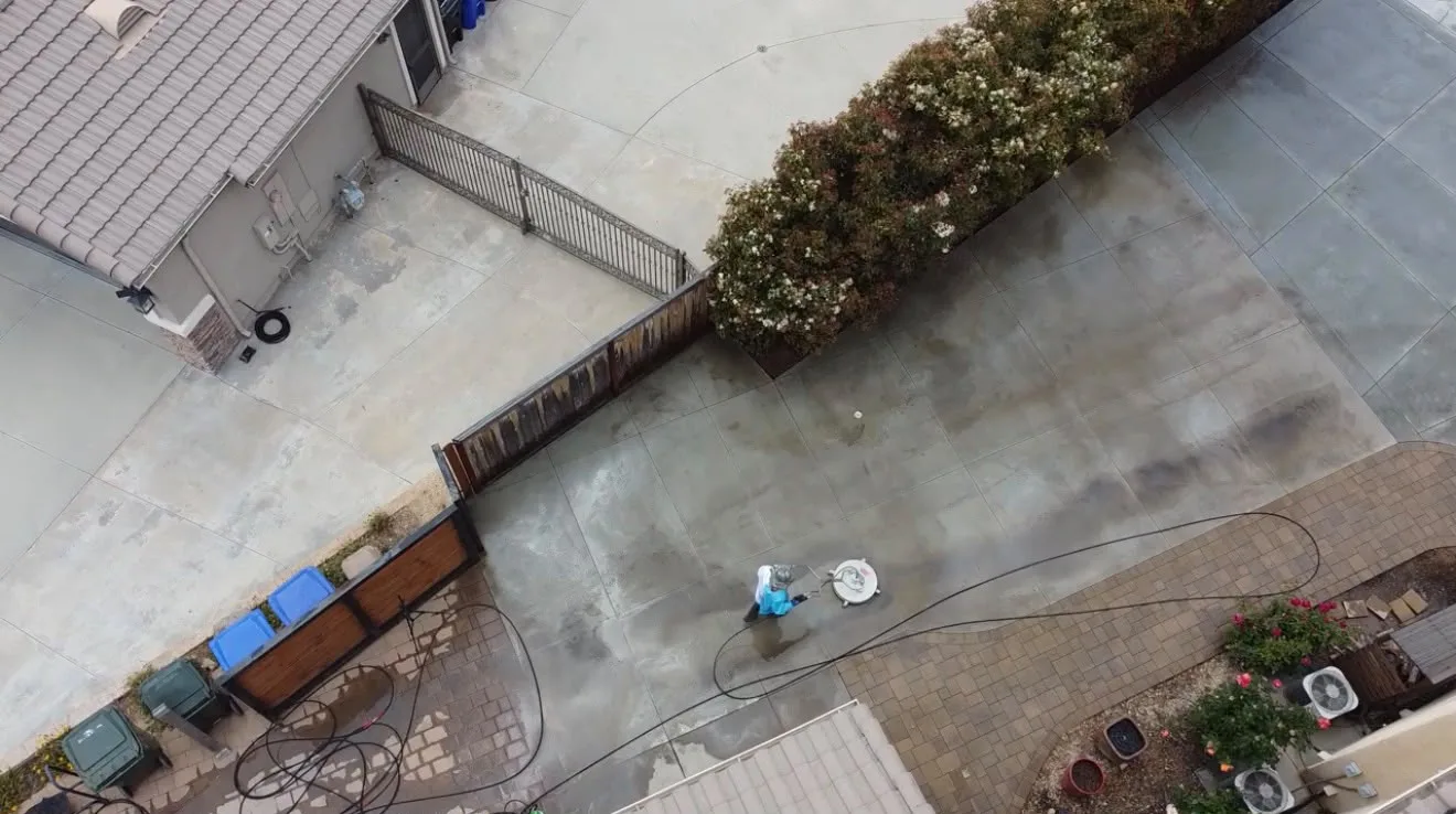 Overhead view of a residential backyard and driveway with a person in blue cleaning near a round utility cover on wet concrete, hoses scattered across the ground, and a fence-lined area with planters and bins nearby.