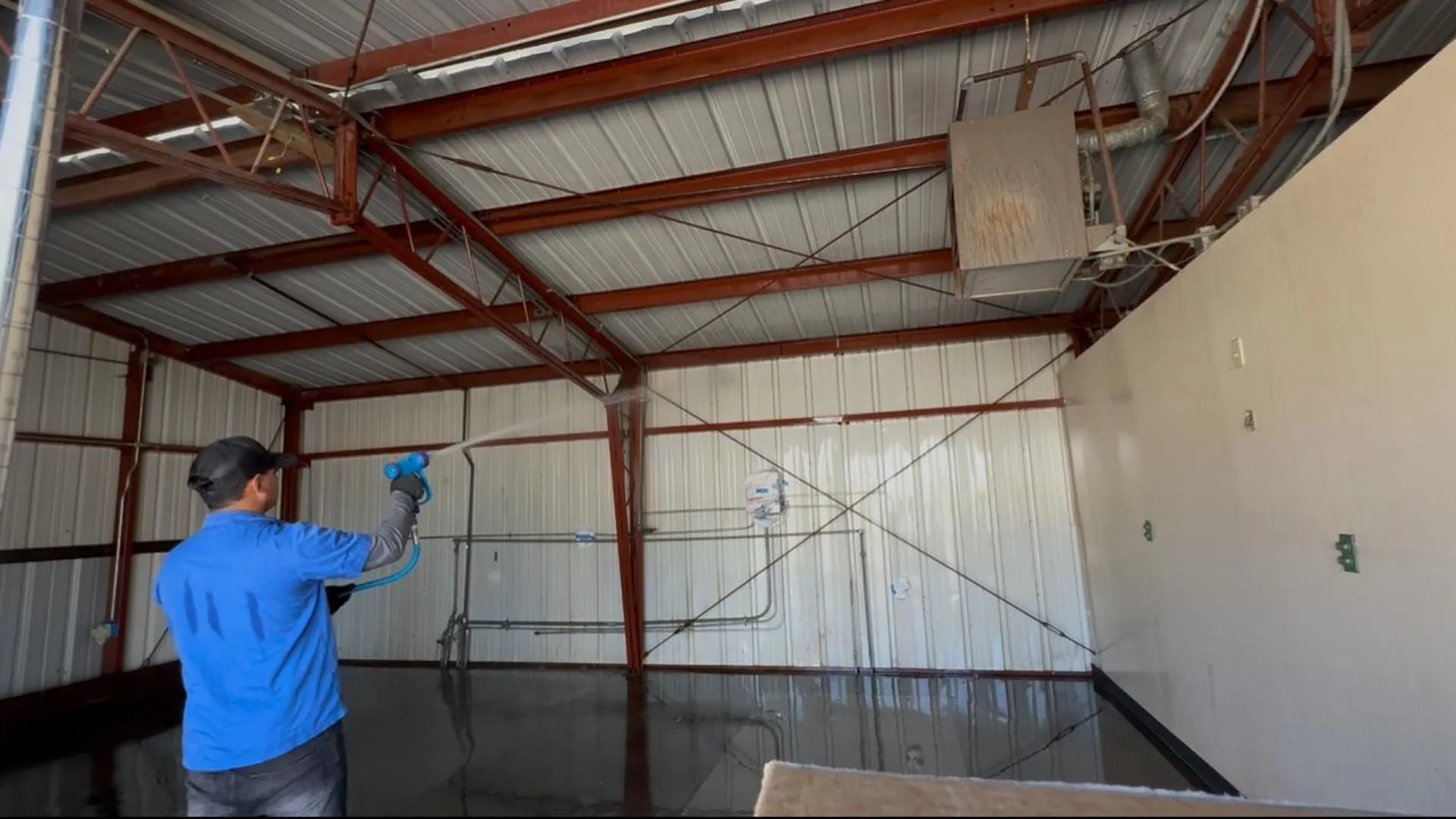 Worker in a blue shirt and cap using a spray wand to clean a wet concrete floor in a metal-walled warehouse with red steel beams overhead.