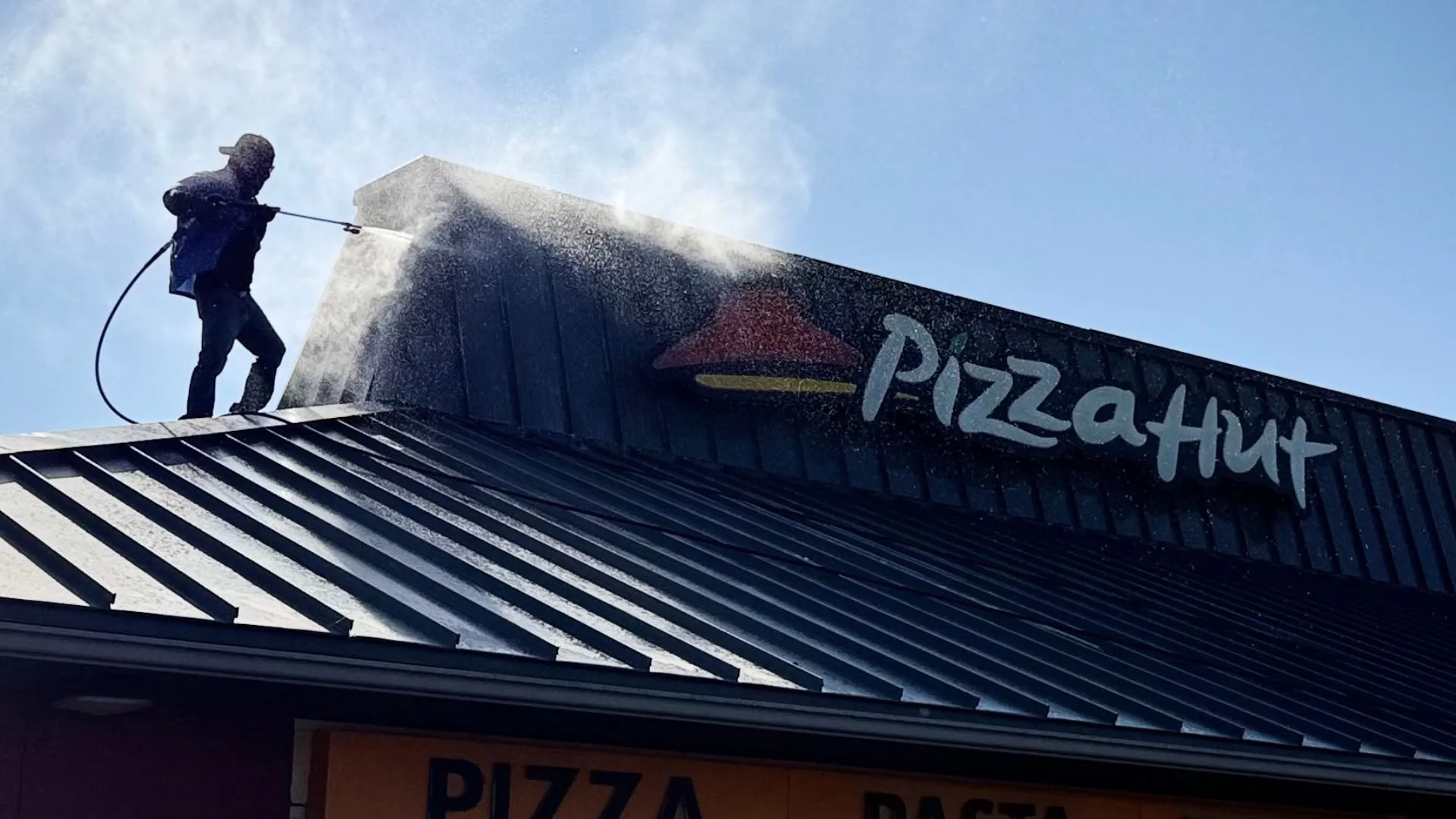 Worker on a roof using a pressure washer to clean the Pizza Hut sign on a sunny day