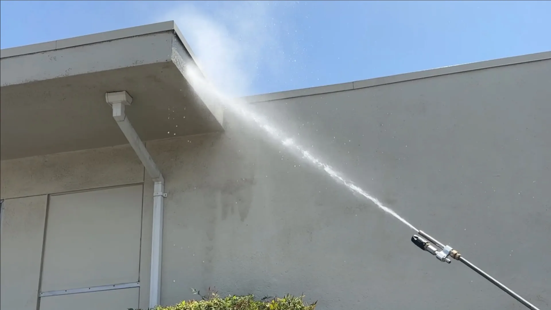 Person using a pressure washer to spray water along the top edge of a building exterior with a high-pressure jet creating mist and spray particles