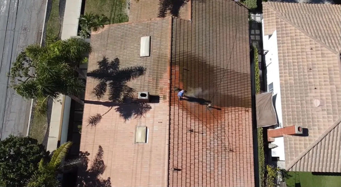 Aerial view of a worker on a red-tiled roof using a hose/pressure washer, with palm tree shadows nearby in a suburban neighborhood.