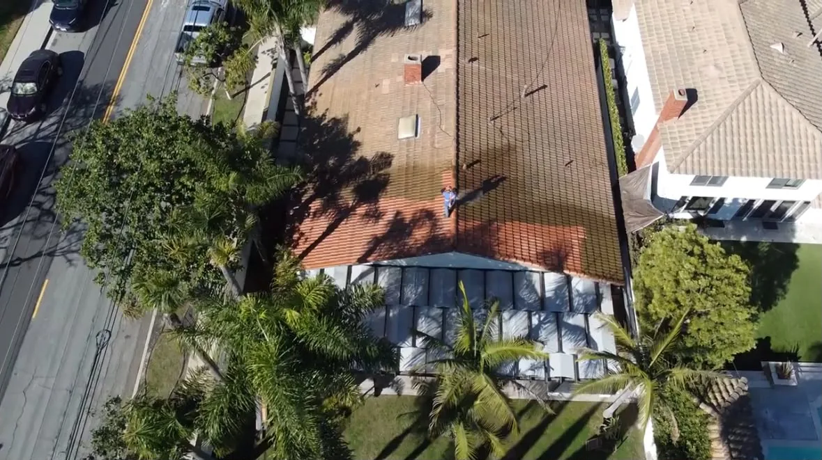 Aerial view of a red-tiled roof house with white awning, surrounded by palm trees and a street with parked cars nearby.