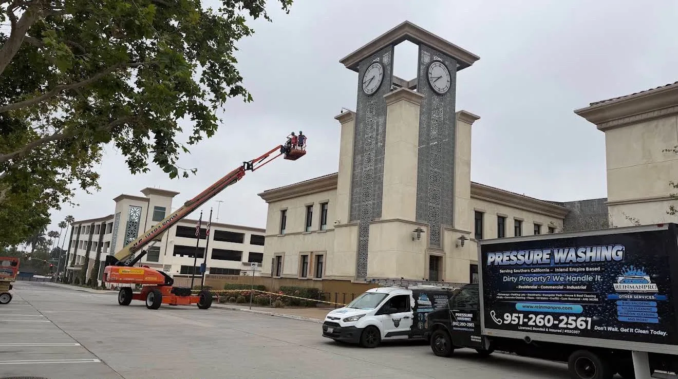 Two workers in a raised bucket wash the clock tower of a beige campus building while a pressure-washing van sits in the parking lot nearby