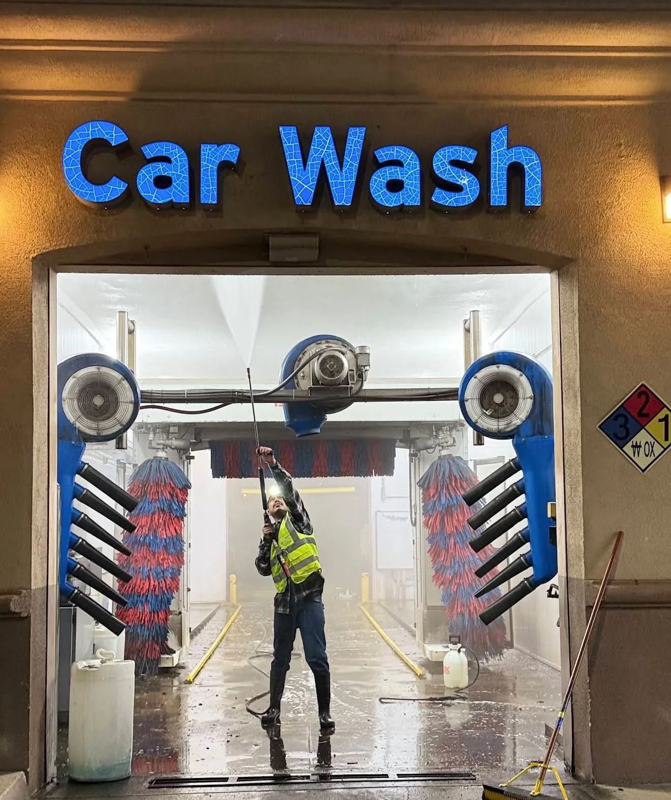 Worker in a neon vest using a spray wand inside a car wash bay with blue rotating brushes nearby at night/indoor lighting