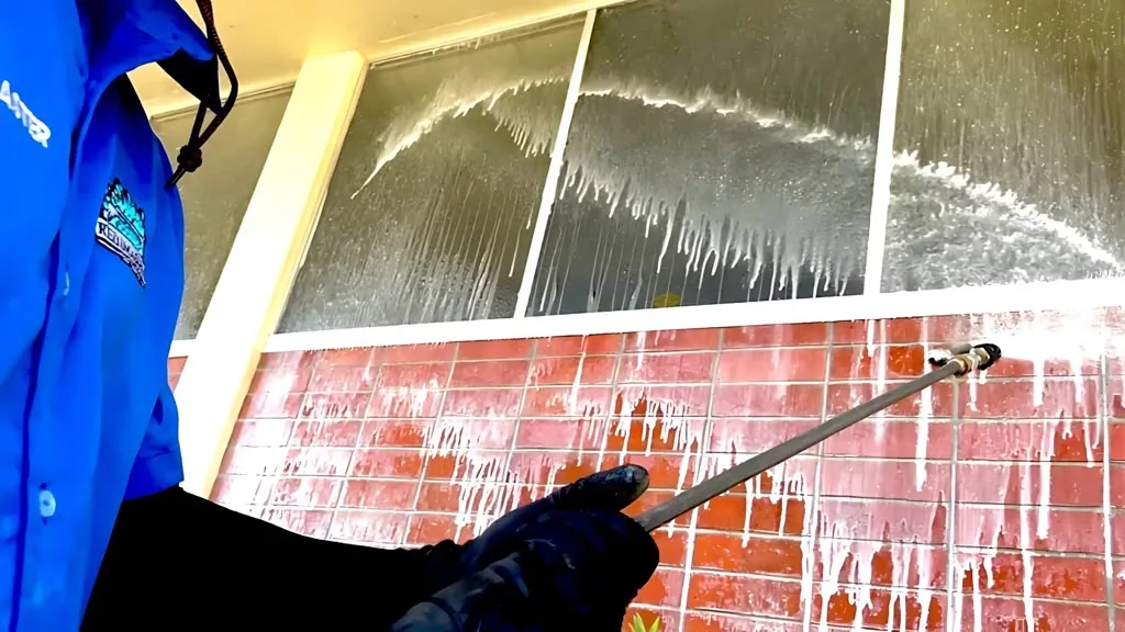 A person in a blue jacket and gloves uses a long spray wand to pressure-wash a red tiled wall and large glass window with soapy water.