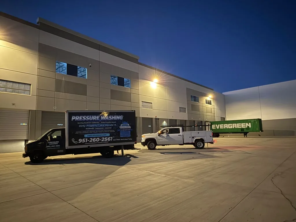 Warehouse loading area at dusk with a black pressure-washing service truck in the foreground and a white service pickup pulling a green Evergreen trailer in the background.