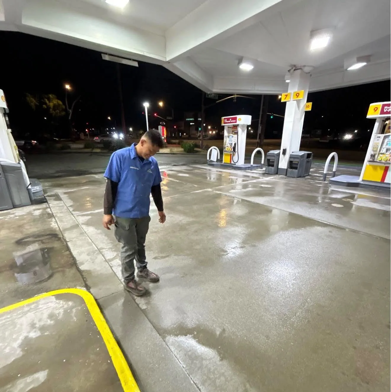 Gas station attendant in blue uniform standing on the wet forecourt under the canopy at night.