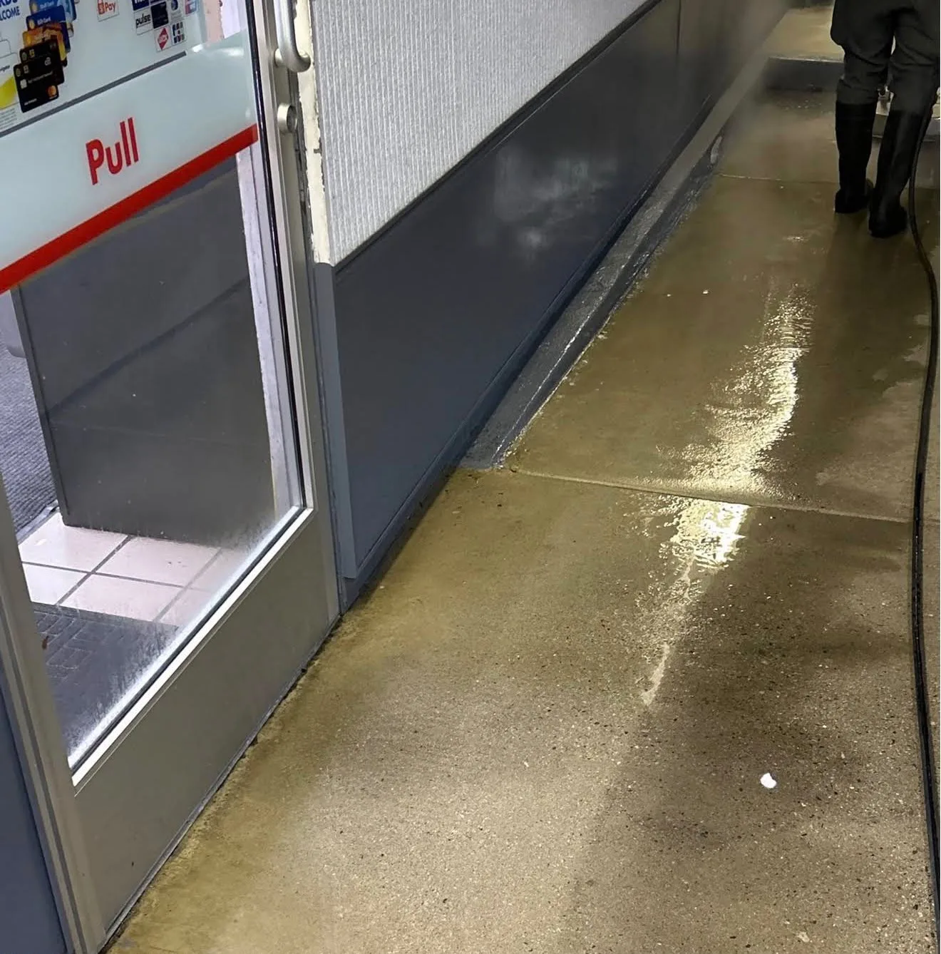 Store entrance with glass door labeled 'Pull'; wet, shiny concrete floor and a person in tall boots visible in the background.