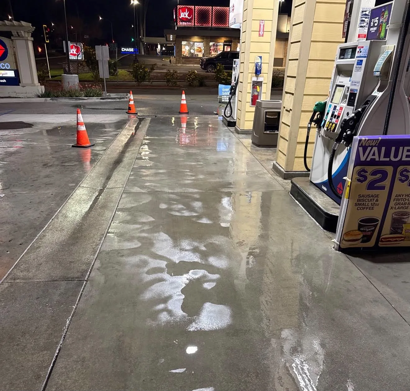 Nighttime gas station scene with wet pavement, reflections on the concrete, and orange traffic cones along the forecourt; fuel pumps visible on the right and storefronts in the distance.