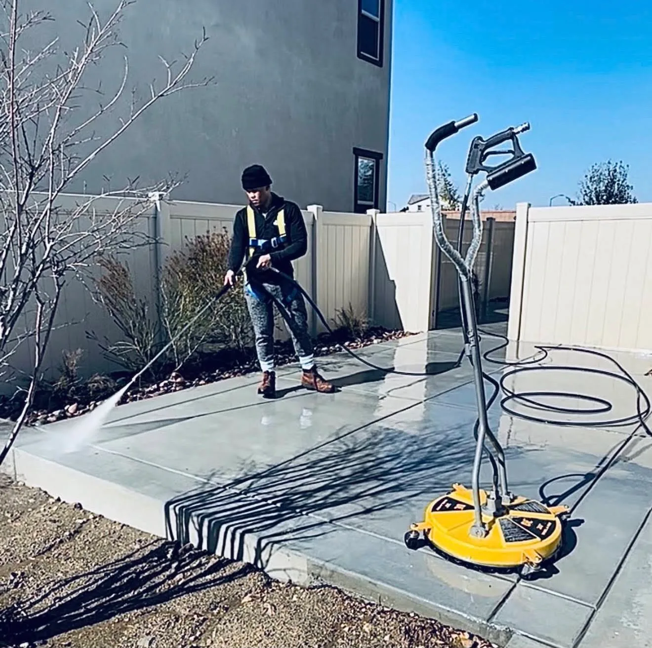 Man in a beanie pressure-washing a concrete patio in a backyard, with a yellow wheeled pressure washer nearby and hoses on the ground.