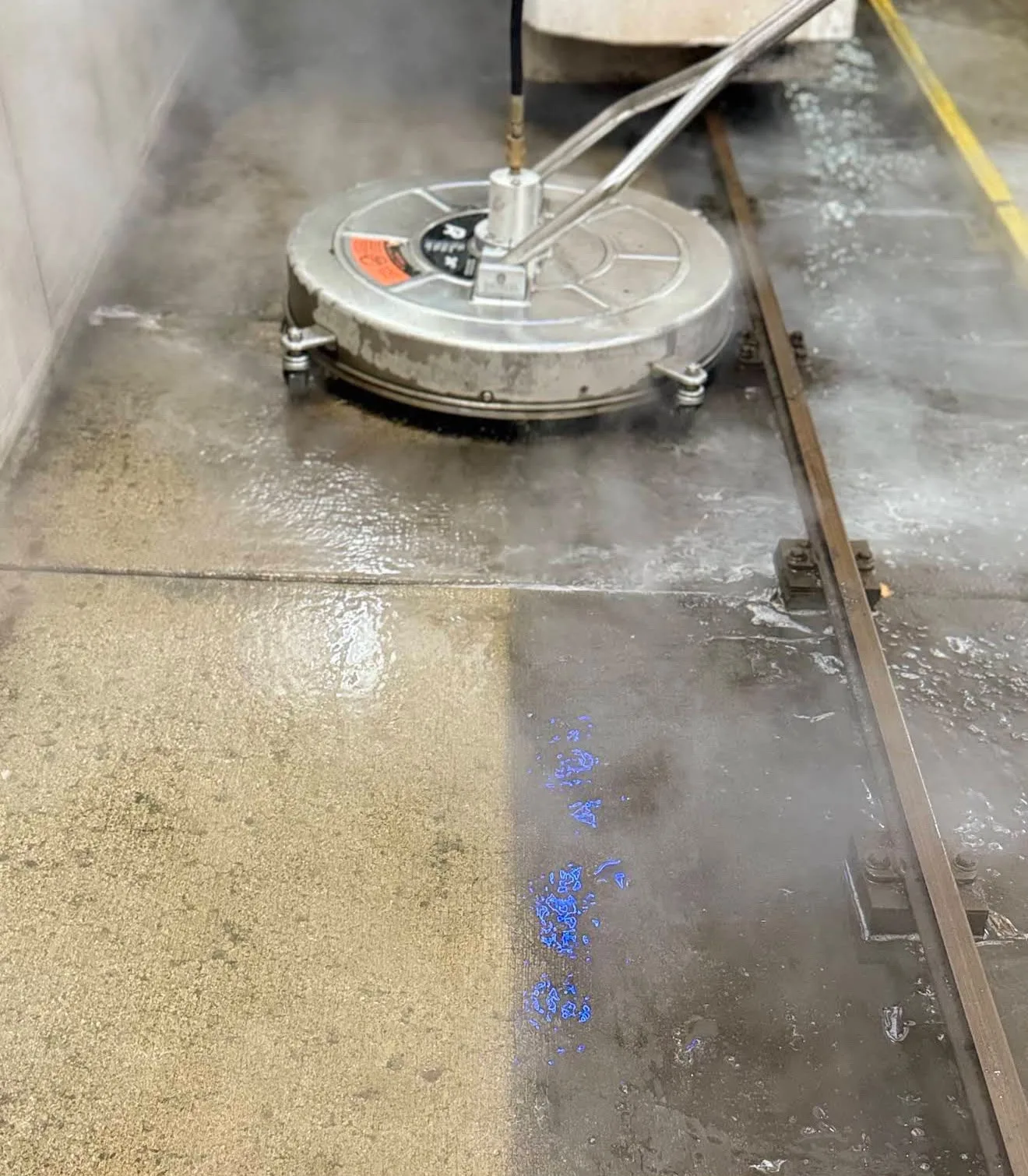 Industrial floor grinding/polishing machine on a wet concrete floor, moving along a metal rail track with blue markings nearby