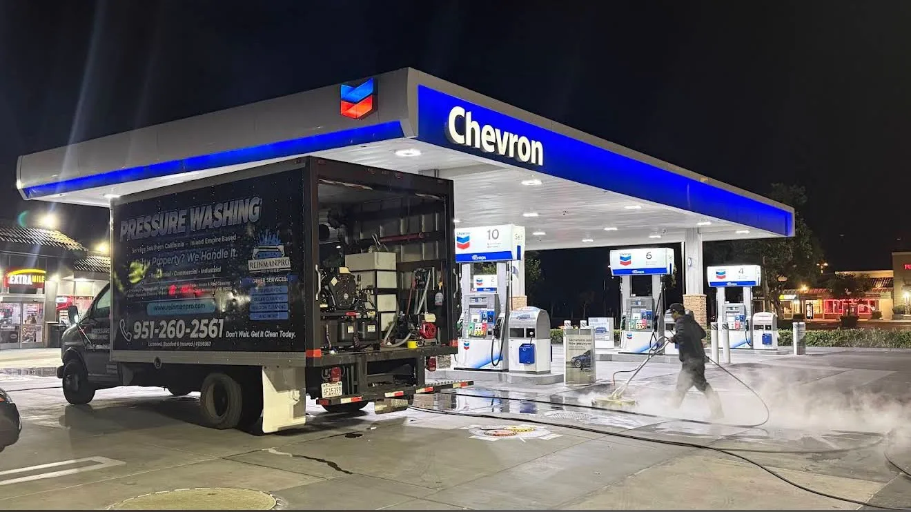 Nighttime Chevron gas station with a pressure-washing truck parked at the pumps while a worker sprays water onto the concrete, creating mist.