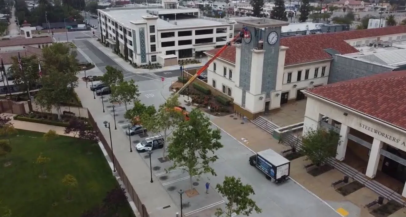 Aerial view of a town square with a clock tower, a lift extending toward the top, and surrounding buildings, trees, and parked cars.