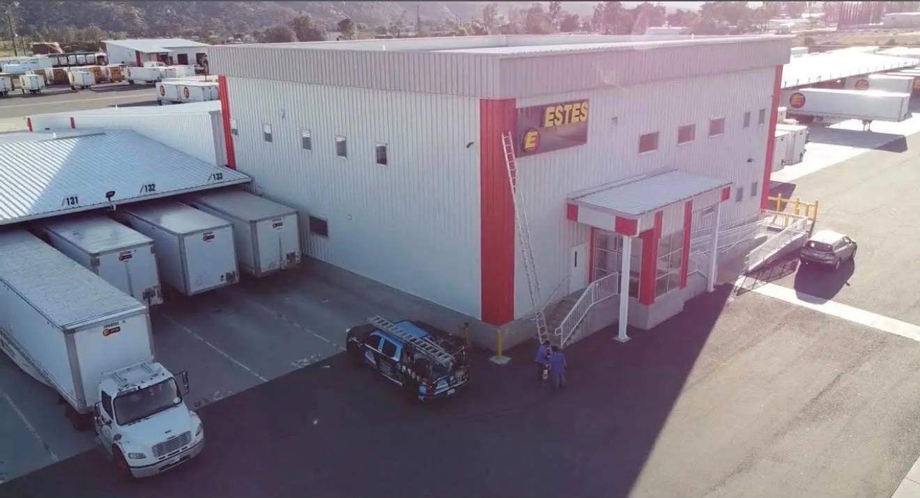 A large industrial loading dock area with delivery trailers parked beside a gray building labeled ESTES; a ladder leans against the wall near the entrance.