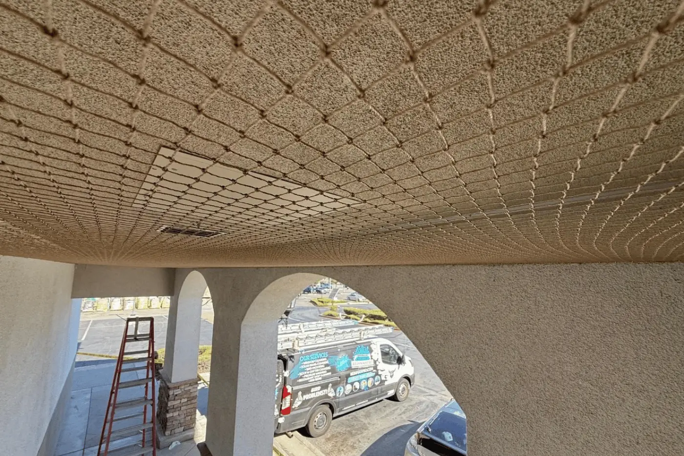 Beige diamond-pattern safety netting across the underside of an overhang, with a red ladder and a service van visible outside.